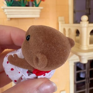 May include: A brown bear figurine wearing a white dress with red polka dots and a red bow. The bear has small black eyes and a fuzzy texture. The figurine is part of a miniature toy set.