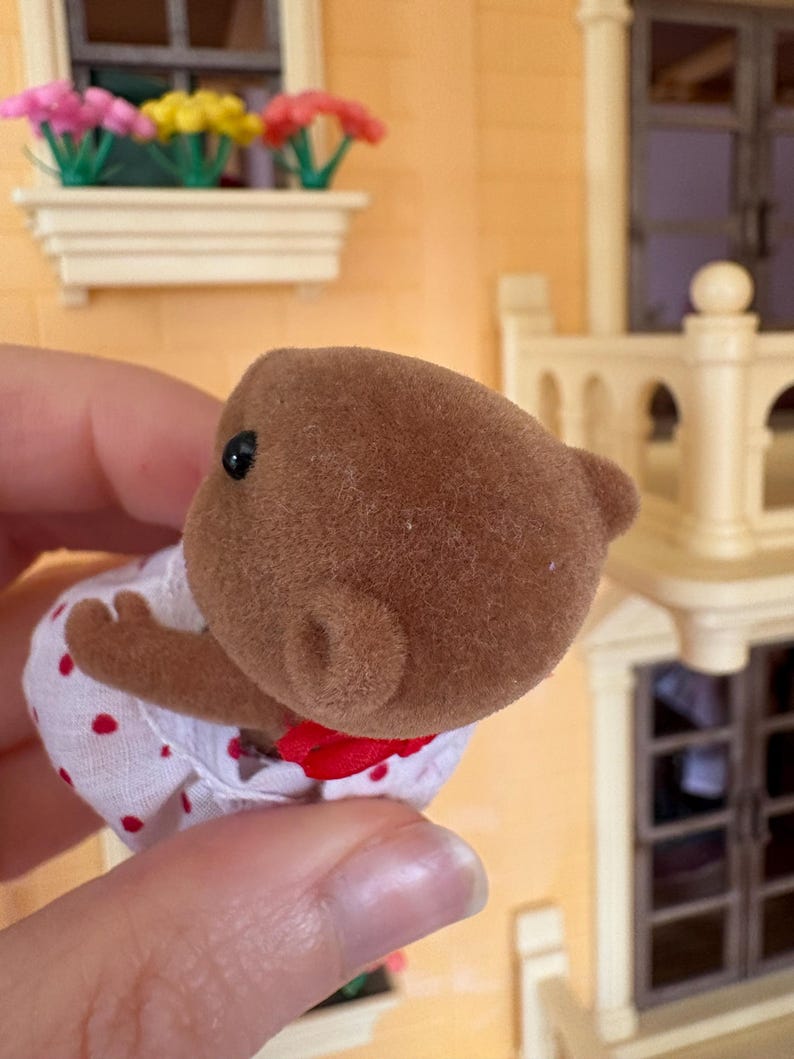 May include: A brown bear figurine wearing a white dress with red polka dots and a red bow. The bear has small black eyes and a fuzzy texture. The figurine is part of a miniature toy set.