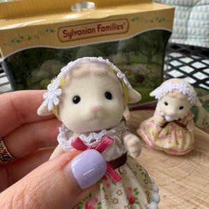 May include: A Sylvanian Families lamb figurine, held in a hand, wearing a floral dress with a pink bow. A second lamb figurine is in the background. The box behind the figurines reads "Sylvanian Families".