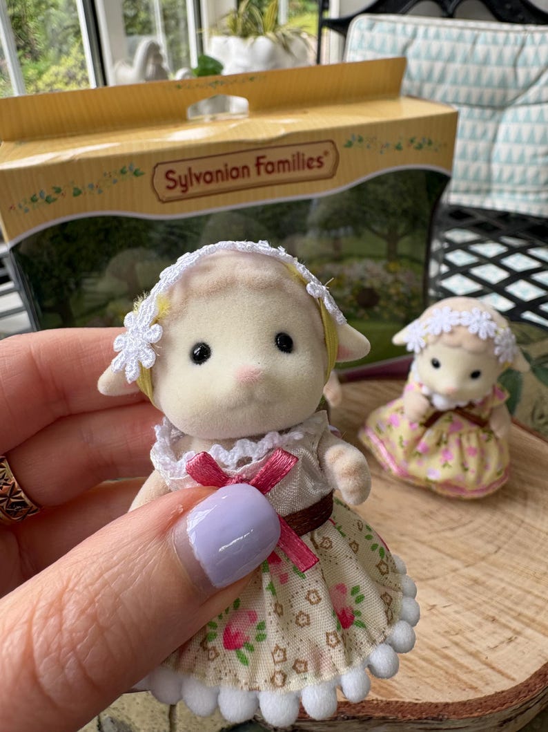 May include: A Sylvanian Families lamb figurine, held in a hand, wearing a floral dress with a pink bow. A second lamb figurine is in the background. The box behind the figurines reads "Sylvanian Families".