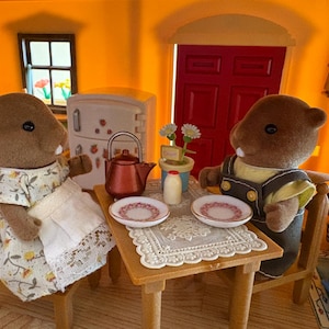May include: A miniature scene featuring two brown beaver figurines seated at a small wooden table. The table is set with plates, a teapot, and a milk bottle. The beavers are dressed in miniature clothing, and the scene is set in a dollhouse.