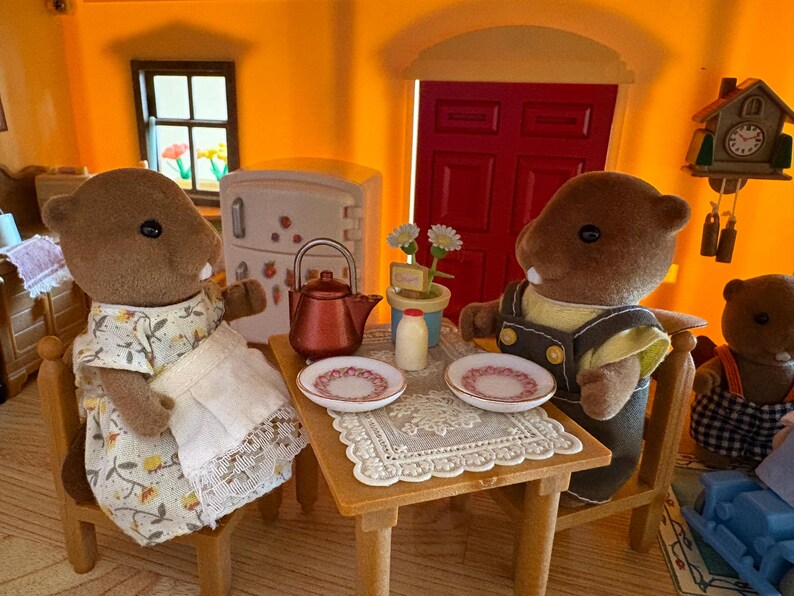 May include: A miniature scene featuring two brown beaver figurines seated at a small wooden table. The table is set with plates, a teapot, and a milk bottle. The beavers are dressed in miniature clothing, and the scene is set in a dollhouse.