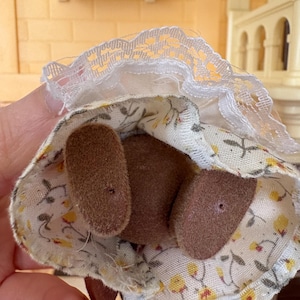 May include: A miniature doll's dress with a floral pattern in yellow and white, paired with brown felt feet. The dress features a white lace trim around the neckline. The doll is part of a miniature toy set.