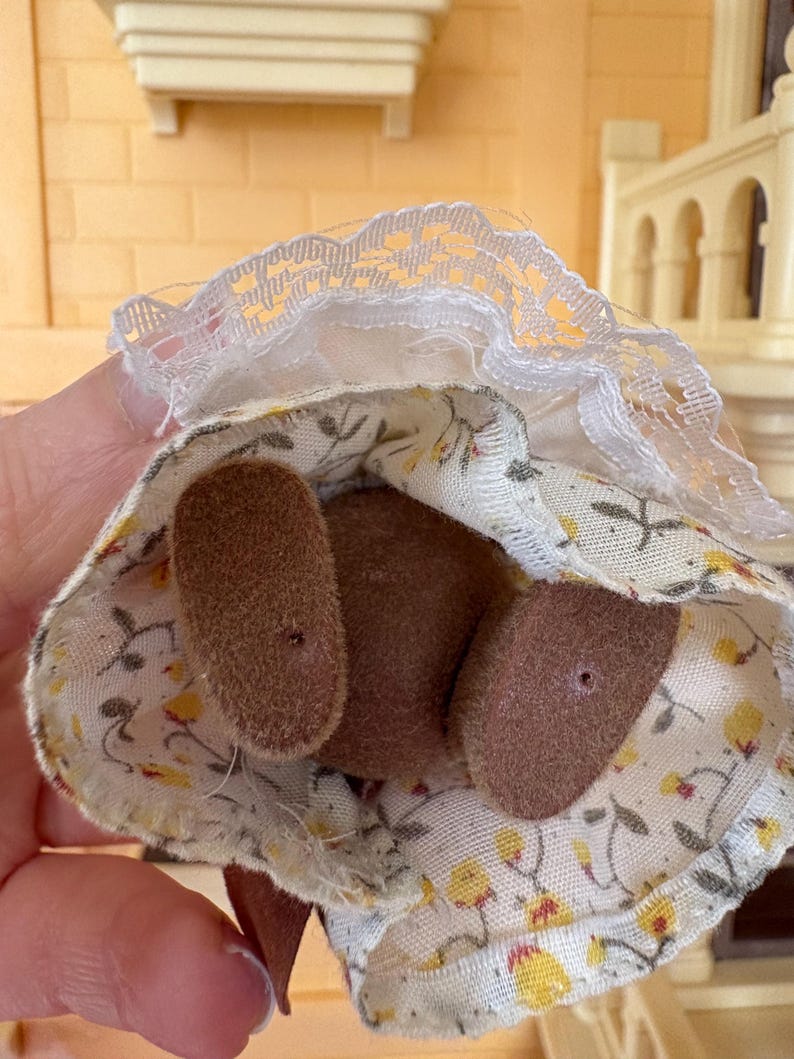May include: A miniature doll's dress with a floral pattern in yellow and white, paired with brown felt feet. The dress features a white lace trim around the neckline. The doll is part of a miniature toy set.