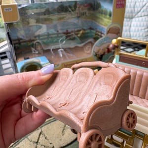 May include: A light brown toy carriage with three seats and four wheels. The carriage is part of a larger toy set, with a box visible in the background featuring a vintage car and a rural scene.