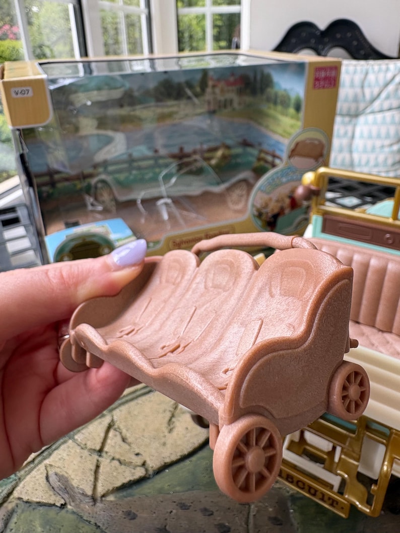 May include: A light brown toy carriage with three seats and four wheels. The carriage is part of a larger toy set, with a box visible in the background featuring a vintage car and a rural scene.