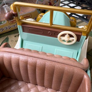 May include: A vintage-style toy car in pastel colors. The car features a light blue body, a brown interior with a quilted pattern, and gold-colored accents. The steering wheel is white. The car has a classic design.