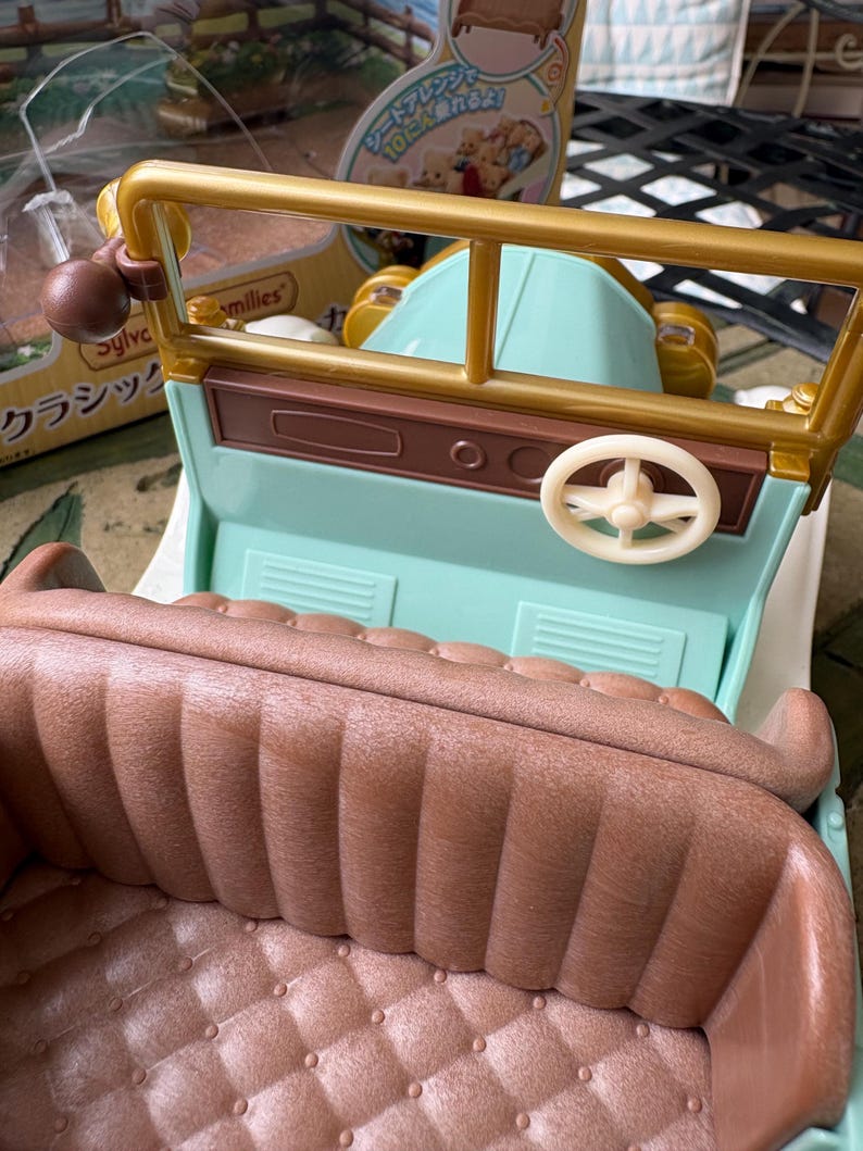 May include: A vintage-style toy car in pastel colors. The car features a light blue body, a brown interior with a quilted pattern, and gold-colored accents. The steering wheel is white. The car has a classic design.