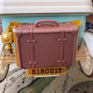 May include: A brown toy suitcase attached to the back of a vintage-style car. The suitcase has a handle and decorative metal accents. The car is light blue and white with gold-colored details. The license plate reads "B15CUIT."