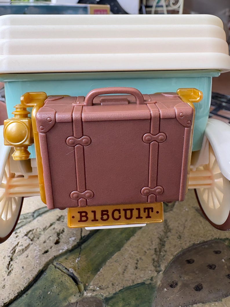 May include: A brown toy suitcase attached to the back of a vintage-style car. The suitcase has a handle and decorative metal accents. The car is light blue and white with gold-colored details. The license plate reads "B15CUIT."