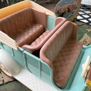 May include: A pastel blue and white toy car with brown seating. The car has a detailed interior with a steering wheel and a decorative exterior. The seats are a quilted brown color. The car is a toy.