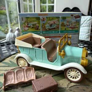 May include: A mint green toy car with cream-colored accents, brown seats, and gold-colored headlights. Includes a brown suitcase and a brown bench. The box behind the car has Japanese text.