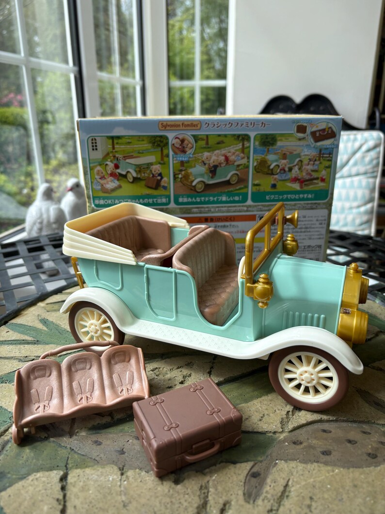 May include: A mint green toy car with cream-colored accents, brown seats, and gold-colored headlights. Includes a brown suitcase and a brown bench. The box behind the car has Japanese text.