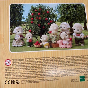 May include: A Sylvanian Families toy set depicting sheep characters in diverse attire, positioned around a tree laden with red apples. The scene includes a pink sofa and a basket, creating a charming miniature world.