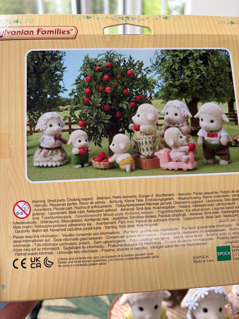 May include: A Sylvanian Families toy set depicting sheep characters in diverse attire, positioned around a tree laden with red apples. The scene includes a pink sofa and a basket, creating a charming miniature world.