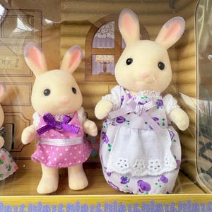 May include: Two toy bunny figures, one in a pink polka-dot dress with a purple bow, the other in a white dress with a purple floral pattern. The bunnies have cream-colored fur and pink-lined ears. The set is in a decorative box.