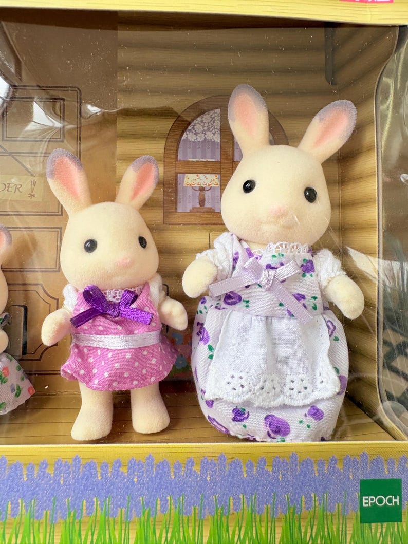 May include: Two toy bunny figures, one in a pink polka-dot dress with a purple bow, the other in a white dress with a purple floral pattern. The bunnies have cream-colored fur and pink-lined ears. The set is in a decorative box.