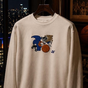 May include: A white crewneck sweatshirt featuring a central graphic of a cartoon character dribbling an orange basketball, with the blue text '26' positioned to the right of the character. The sweatshirt has long sleeves and ribbed cuffs.