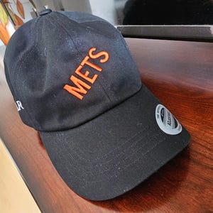 May include: Black baseball cap with the word "METS" in orange block letters on the front. The cap has a curved brim and an adjustable strap. A small white label is visible on the side.