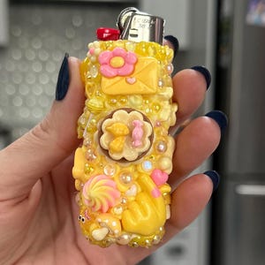 May include: A yellow lighter decorated with various embellishments. The lighter features a pink flower, a yellow envelope, and a swirl design. The lighter is covered in small, clear beads and other decorative elements.