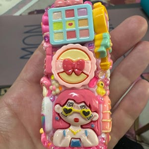 May include: A pink, decorative, rectangular object with a cartoon girl design. The object features a makeup palette, a compact with a bow, and a girl with heart-shaped sunglasses. The object is adorned with rhinestones and other decorative elements.