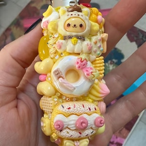May include: A decorative lighter with a whimsical design. The lighter is covered in yellow and white sculpted elements, including a cat character, a donut, and a cake. Pink and brown accents add detail to the design.