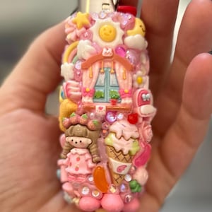 May include: A pink lighter decorated with various miniature charms, including a smiling sun, rainbow, ice cream cone, and a doll. The lighter is embellished with sparkling rhinestones and other decorative elements.