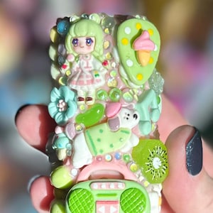 May include: A decorative, rectangular object with a variety of colorful, miniature embellishments. Features include a doll, a dog, a kiwi slice, and a boombox. The object is encrusted with small, clear rhinestones.