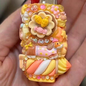 May include: A decorative lighter with a whimsical design. The lighter is covered in colorful, sculpted elements including a flower, a dog in a pink sweater, and candy-like embellishments. The lighter is held in a hand.