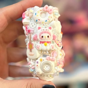 May include: A white, decorative object adorned with various miniature elements. It features a donut with a pink bow, a cat-like character, a camera, and a guitar. The object is embellished with rhinestones and other small, colorful details.