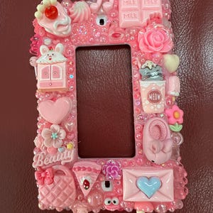 May include: Pink decorative light switch cover with various embellishments. Features a cake, a bunny, a cat, a shoe, and a basket. The word "Beauty" is written on the cover. The cover is surrounded by small, clear beads.