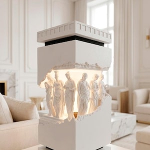 Könnte beinhalten: Eine weiße Tischlampe im architektonischen Stil mit skulptierten Figuren. Die Lampe hat einen rechteckigen Sockel und eine mehrschichtige Oberseite, wobei ein Teil ausgeschnitten ist, um die beleuchteten Figuren freizulegen. Die Lampe steht auf einem weißen Tisch.
