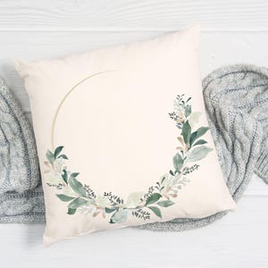 May include: Cream-colored square pillow with a watercolor floral wreath design. The wreath features green and white leaves and a gold-colored arc. A gray knitted blanket is draped on either side of the pillow.