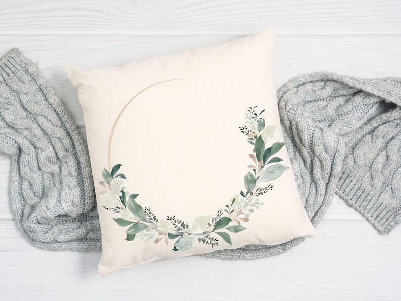 May include: Cream-colored square pillow with a watercolor floral wreath design. The wreath features green and white leaves and a gold-colored arc. A gray knitted blanket is draped on either side of the pillow.