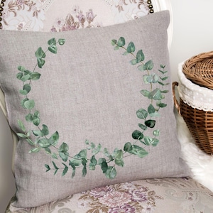 May include: A decorative gray pillow with a watercolor eucalyptus wreath design. The pillow is on a floral patterned chair, with a wicker basket in the background. The eucalyptus leaves are green.