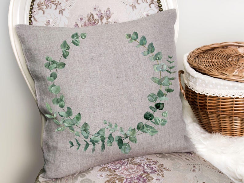 May include: A decorative gray pillow with a watercolor eucalyptus wreath design. The pillow is on a floral patterned chair, with a wicker basket in the background. The eucalyptus leaves are green.