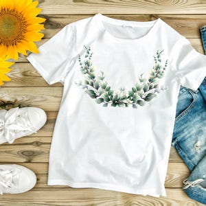May include: White t-shirt with a watercolor eucalyptus leaf design. The shirt is paired with blue denim jeans and white sneakers. Two sunflowers are placed on a wooden surface.