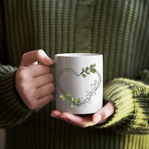 May include: White ceramic mug featuring a black heart outline with green leaf accents. The mug is held by a person wearing a green sweater. The design is simple and elegant, suitable for beverages.