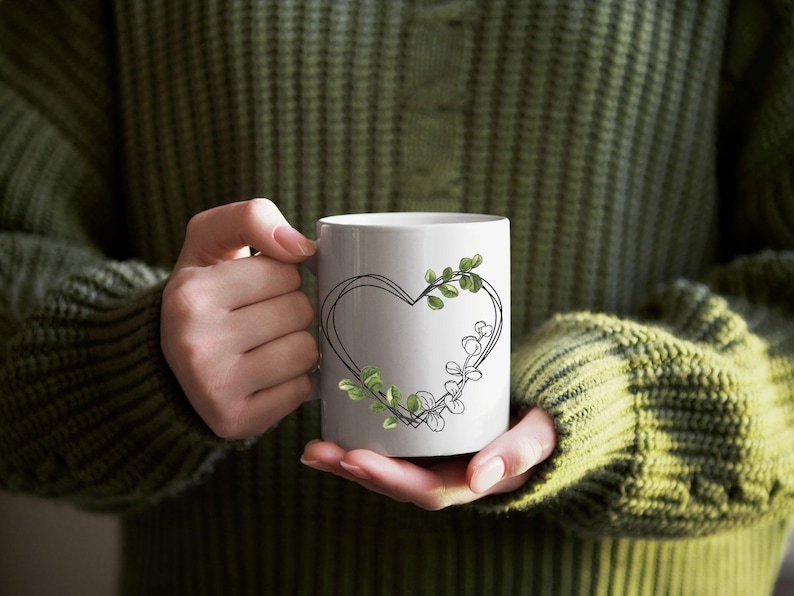 May include: White ceramic mug featuring a black heart outline with green leaf accents. The mug is held by a person wearing a green sweater. The design is simple and elegant, suitable for beverages.