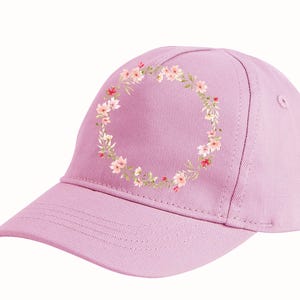 May include: A light purple baseball cap with a floral wreath design. The wreath is composed of small pink and red flowers with green leaves. The cap has a curved brim and a structured crown.