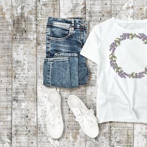 May include: A flat lay featuring a white t-shirt with a purple floral wreath design, folded blue denim jeans, and white lace-up sneakers. The items are arranged on a weathered, light-colored wooden surface.