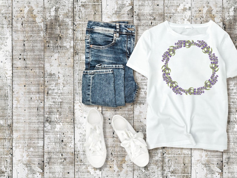 May include: A flat lay featuring a white t-shirt with a purple floral wreath design, folded blue denim jeans, and white lace-up sneakers. The items are arranged on a weathered, light-colored wooden surface.