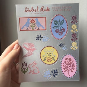 May include: A sheet of stickers with cross-stitch designs. The stickers feature birds, flowers, and other motifs in pink, blue, yellow, and red. The text "isobel Rose ILLUSTRATION CROSS STITCH COLLECTION" is printed at the top.
