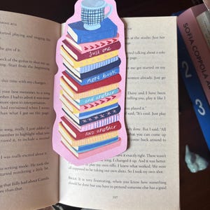 Puede incluir: Un marcapáginas caprichoso con una pila de libros coloridos con las frases "Just one", "more book" y "and another". Una taza azul y blanca se encuentra encima de la pila de libros. El marcapáginas está en una página de libro.