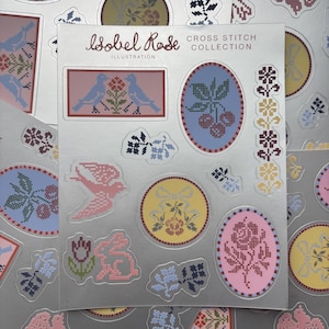 May include: A sticker sheet titled Isobel Rose Cross Stitch Collection featuring various die-cut designs including blue birds, cherries, a pink bird, a yellow circle with a bow and flower, a pink oval with a rose, and floral border elements.