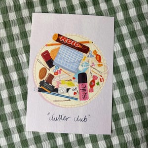 Puede incluir: Una impresión que presenta una colección de artículos cotidianos dispuestos sobre una superficie circular. Los artículos incluyen una barra de chocolate Rolo, una pequeña caja azul, esmalte de uñas y un reloj. La impresión se titula "clutter club".