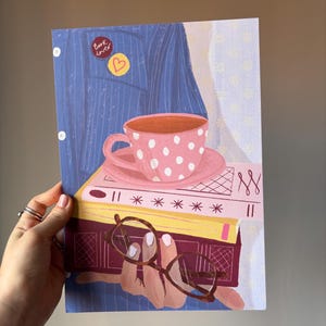 May include: An illustration of a blue shirt with two pins, one reading Book lover and one with a heart. A pink polka dot teacup sits on a stack of three books. A hand holds a pair of brown glasses in front of the books, which are pink, yellow, and dark red.