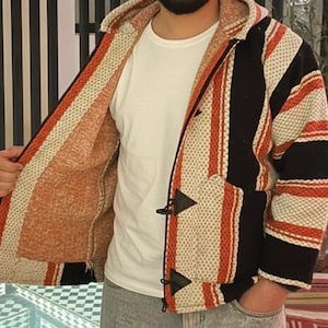 Op de afbeelding: Een gestreepte jas met capuchon in zwart, crème en oranje. De jas heeft een knevelsluiting en een getextureerde afwerking. De binnenvoering is effen oranje. Er wordt een wit T-shirt onder gedragen.