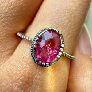 May include: A silver ring featuring an oval, faceted, pink gemstone. The gemstone is set in a beaded bezel, and the band has a twisted rope design. The ring is shown on a finger.
