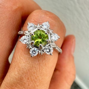 May include: A silver ring featuring a central, round, light green gemstone surrounded by white, sparkling accent stones. The ring's design includes a floral-like arrangement of the accent stones, creating a halo effect around the main stone. The ring is on a finger.